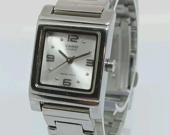 Könnte beinhalten: Eine silberfarbene Casio Quartz-Uhr mit quadratischem Zifferblatt und passendem Metallarmband. Das Zifferblatt ist silberfarben mit schwarzen Ziffern und Zeigern. Die Uhr zeigt die Uhrzeit und den Markennamen an.