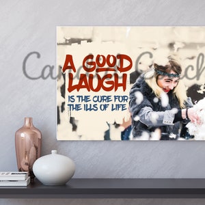 May include: A white canvas print with a red and blue graphic that says "A good laugh is the cure for the ills of life". A woman is smiling and throwing snow in the background.