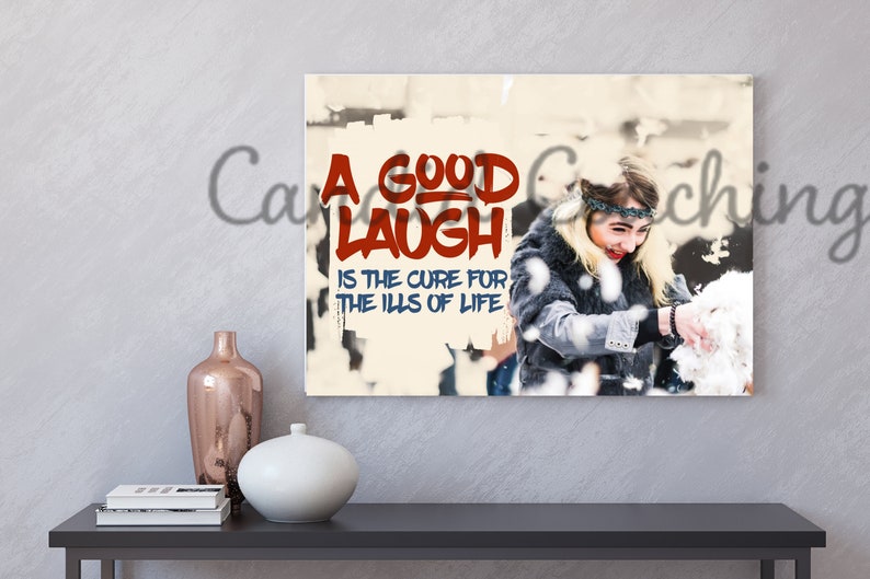 May include: A white canvas print with a red and blue graphic that says "A good laugh is the cure for the ills of life". A woman is smiling and throwing snow in the background.
