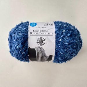 May include: A skein of blue and white yarn called "Cozy Boucle" by Loops & Threads. The yarn is a soft, fluffy boucle style and is perfect for knitting or crocheting blankets, scarves, and other cozy items.