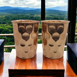 May include: Two silver cups with a hammered texture sit on a wooden surface in front of a window with a view of a green valley and mountains in the distance.