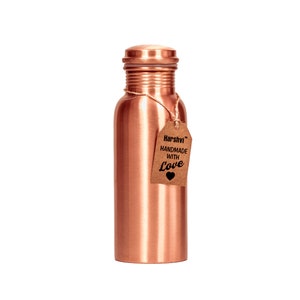 May include: A cylindrical copper water bottle with a smooth metallic finish and a matching screw-top lid. A brown paper tag is tied to the neck with twine, featuring the text Harshvi HANDMADE WITH Love and a small black heart icon printed on it.