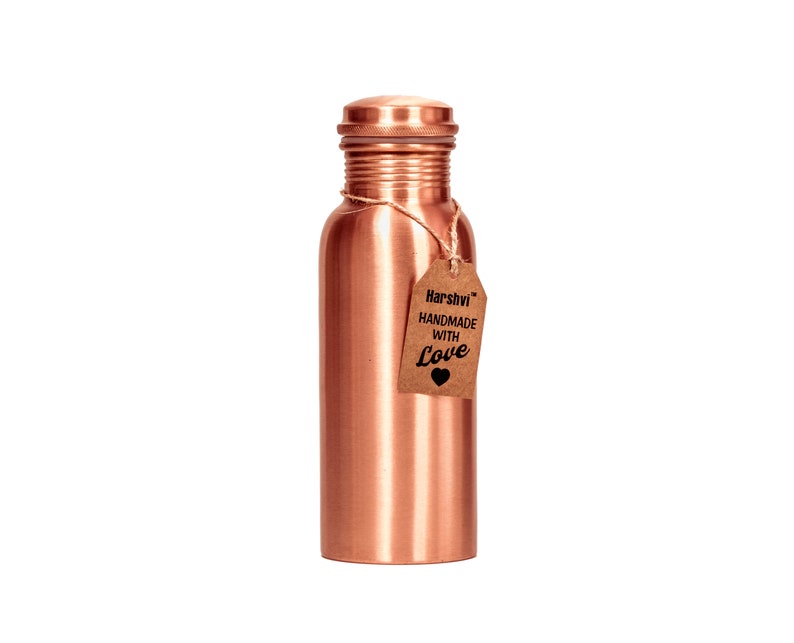 May include: A cylindrical copper water bottle with a smooth metallic finish and a matching screw-top lid. A brown paper tag is tied to the neck with twine, featuring the text Harshvi HANDMADE WITH Love and a small black heart icon printed on it.