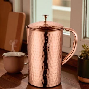 May include: A copper-colored hammered metal French press coffee maker with a lid and handle. A matching mug sits nearby, with steam rising from it. A small potted plant is to the right.