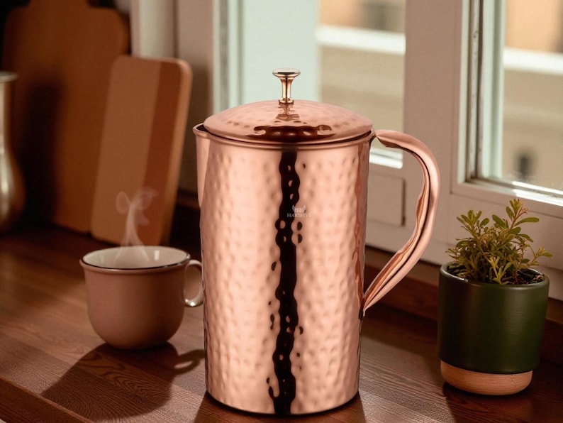 May include: A copper-colored hammered metal French press coffee maker with a lid and handle. A matching mug sits nearby, with steam rising from it. A small potted plant is to the right.