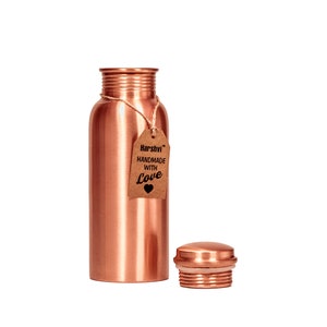 May include: A cylindrical copper water bottle with a metallic finish and a threaded neck. A brown paper tag is tied to the neck with twine, featuring the text "Harshvi HANDMADE WITH Love" and a heart icon. A matching copper screw-on cap sits nearby.