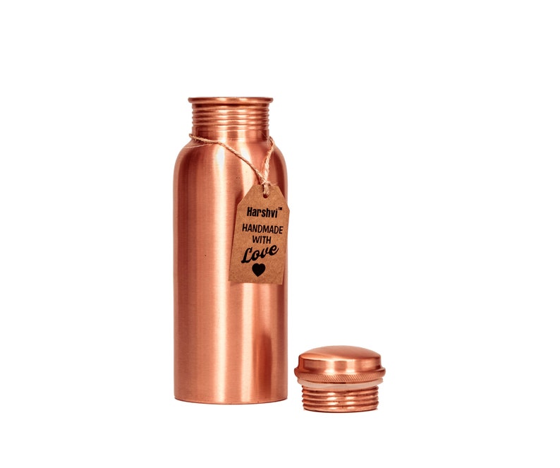 May include: A cylindrical copper water bottle with a metallic finish and a threaded neck. A brown paper tag is tied to the neck with twine, featuring the text "Harshvi HANDMADE WITH Love" and a heart icon. A matching copper screw-on cap sits nearby.
