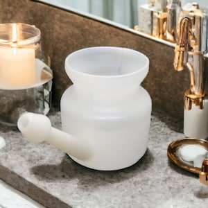 Op de afbeelding: Witte keramische neti pot met een tuit op een grijs aanrecht. De neti pot wordt gebruikt voor neusdouchen.