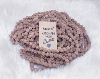 Puede incluir: Primer plano de un collar de cuentas mala marrón hecho de cuentas de madera natural. El collar está sobre una superficie blanca y peluda. Una pequeña etiqueta de papel marrón con el texto "Harshvi™ Hecho a mano con amor" es visible.
