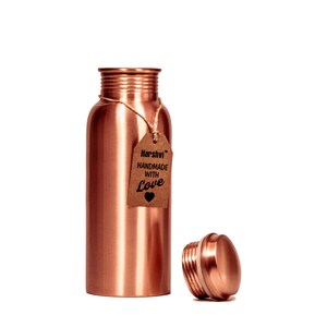 May include: A cylindrical copper water bottle with a smooth, metallic finish and a threaded neck. A brown paper tag is tied to the neck with twine, featuring the text Harshvi HANDMADE WITH Love and a small heart icon. The matching cap sits nearby.