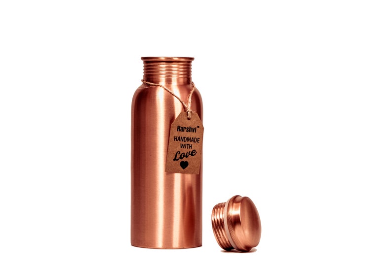 May include: A cylindrical copper water bottle with a smooth, metallic finish and a threaded neck. A brown paper tag is tied to the neck with twine, featuring the text Harshvi HANDMADE WITH Love and a small heart icon. The matching cap sits nearby.
