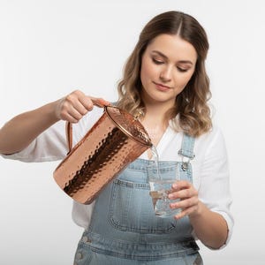 May include: A copper pitcher with a hammered texture is pouring water into a clear glass. The pitcher has a handle and a lid. The person is wearing a white shirt and denim overalls.