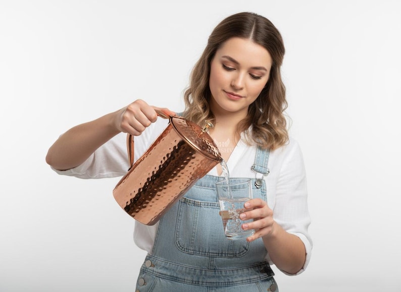 May include: A copper pitcher with a hammered texture is pouring water into a clear glass. The pitcher has a handle and a lid. The person is wearing a white shirt and denim overalls.