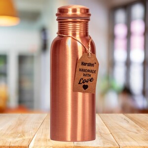 May include: A cylindrical copper water bottle with a smooth metallic finish and a matching screw-top lid. A brown paper tag is tied around the neck with twine, featuring the text Harshvi HANDMADE WITH Love and a small black heart icon.