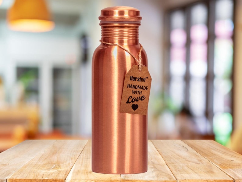 May include: A cylindrical copper water bottle with a smooth metallic finish and a matching screw-top lid. A brown paper tag is tied around the neck with twine, featuring the text Harshvi HANDMADE WITH Love and a small black heart icon.