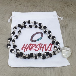 Puede incluir: Un collar de cuentas negras con una cadena de plata y un colgante de concha blanca. El collar está sobre una bolsa de tela blanca con la palabra "HARSHVI" impresa en ella.