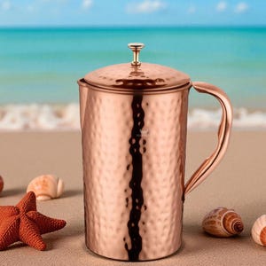 May include: A copper pitcher with a hammered texture, a lid, and a curved handle. The pitcher is on a sandy beach with seashells and a starfish. The ocean is in the background.
