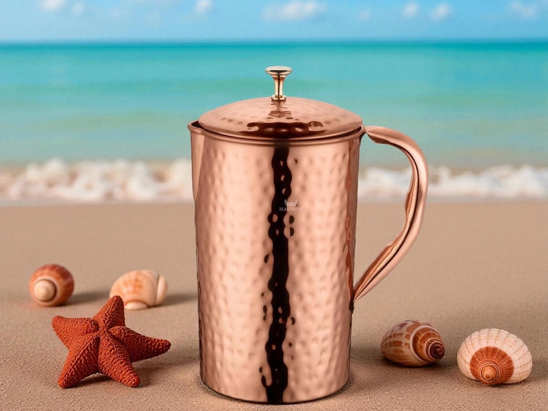 May include: A copper pitcher with a hammered texture, a lid, and a curved handle. The pitcher is on a sandy beach with seashells and a starfish. The ocean is in the background.