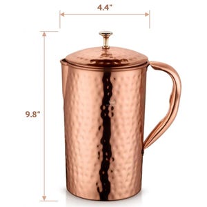 May include: A copper pitcher with a hammered texture, a lid, and a curved handle. The pitcher is 9.8 inches tall and 4.4 inches wide. The pitcher has a shiny, metallic finish and is designed for serving beverages.
