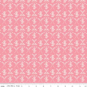 May include: Pink fabric with a repeating pattern of off-white floral and bow designs. The pattern is symmetrical and covers the entire surface. The fabric has a ruler along the bottom edge, with the text "Inches" and "Riley Blake Designs".