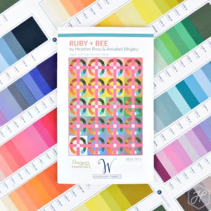 May include: A fabric pattern book titled "RUBY + BEE" by Heather Ross & Annabel Wrigley, featuring a colorful geometric design. The book is surrounded by fabric swatches in various colors, including pink, blue, green, and yellow. The text "Designer Essentials" and "WINDHAM FABRICS" are visible.