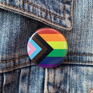 May include: A round button with a rainbow pride flag design. The flag includes the colors of the transgender pride flag, the black and brown stripes of the progress pride flag, and the traditional rainbow pride flag colors.