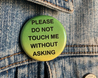 以下が含まれることがあります： 「PLEASE DO NOT TOUCH ME WITHOUT ASKING」の黒い文字が入った、丸い緑と黄色のボタン。青いデニムジャケットにピンで留められています。ジャケットにはステッチと銀色のボタンが見えます。