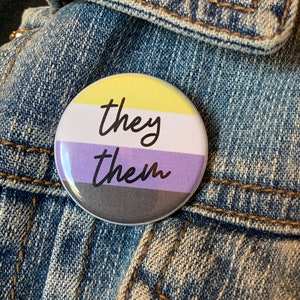 May include: A yellow, purple and black button with the text "they them" in black.