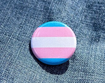 May include: A blue, white, and pink transgender pride button pin on a blue denim background.