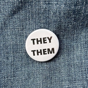 May include: White button with the text "THEY THEM" on a blue denim background.