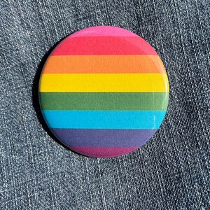 May include: A round button with a rainbow design. The button has six stripes in the colors of the rainbow: red, orange, yellow, green, blue, and purple.