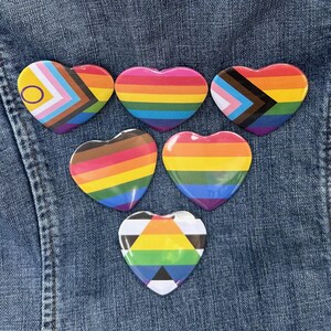 May include: Six heart-shaped pins with rainbow and LGBTQ+ pride flag designs. The pins are arranged on a blue denim background. Each pin features a different flag design, including the Progress Pride flag and the Pride flag.