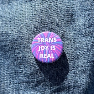 May include: A purple button with a pink and blue firework design. The button says "Trans Joy is Real" in white text.