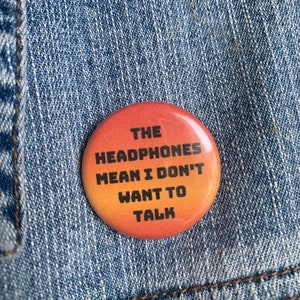 May include: A round button with a gradient background of orange, yellow, and red. The button has black text that reads "THE HEADPHONES MEAN I DON'T WANT TO TALK".