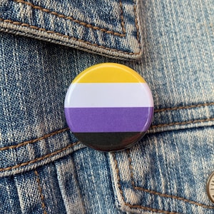 May include: A yellow, white, purple, and black button with the colors arranged in horizontal stripes. The button is pinned to a blue denim jacket.