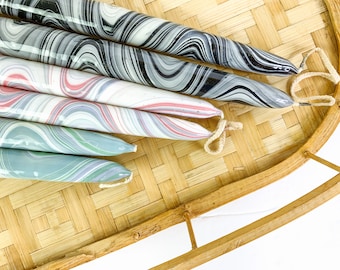 May include: Six taper candles with a marbled design in shades of black, gray, pink, white, and blue. The candles are arranged on a woven bamboo tray with a light-colored background. Each candle has a small loop of twine.