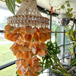 May include: A tiered hanging mobile crafted from numerous orange and cream-colored scallop shells. The structure features a top section of woven white cord and small shells, with multiple cascading layers of orange shells suspended by thin white strings.