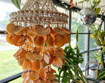 May include: A tiered hanging mobile crafted from numerous orange and cream-colored scallop shells. The structure features a top section of woven white cord and small shells, with multiple cascading layers of orange shells suspended by thin white strings.