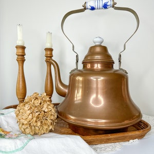 May include: A copper tea kettle with a white porcelain handle and lid, sitting on a woven tray with two wooden candle holders and a dried hydrangea.