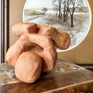 May include: A terracotta-colored abstract sculpture with rounded forms. The sculpture sits on a vintage book with a leather cover. In the background, a framed landscape painting depicts trees and a field.