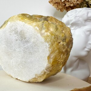 May include: A large, irregularly shaped stone with a flat, white, polished surface and a rough, textured exterior in shades of yellow and tan. The stone is displayed near a white bust and dried flowers.