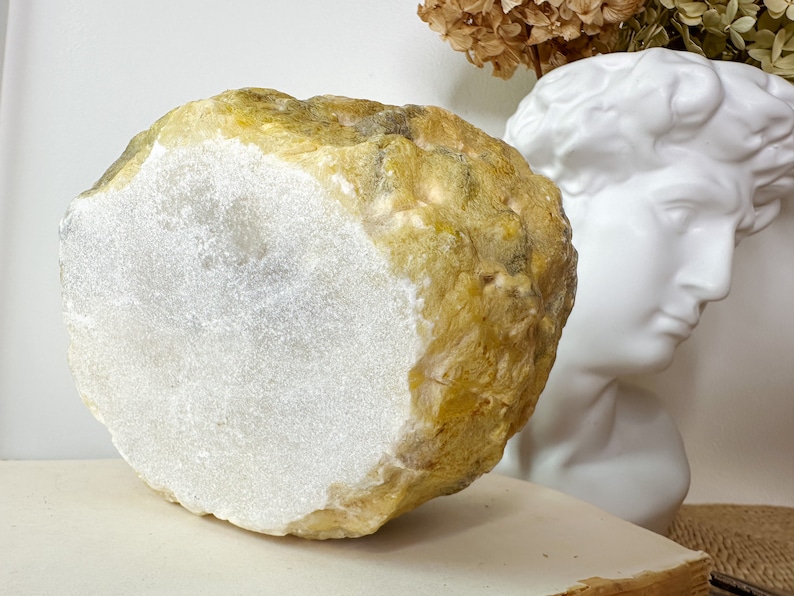 May include: A large, irregularly shaped stone with a flat, white, polished surface and a rough, textured exterior in shades of yellow and tan. The stone is displayed near a white bust and dried flowers.