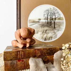May include: A terracotta-colored sculpture of a person in a seated, contemplative pose. The sculpture rests on two antique books with gold lettering. A framed landscape art piece with trees and a field is in the background.