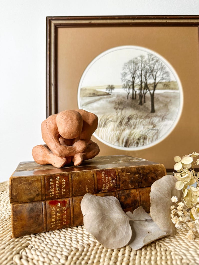 May include: A terracotta-colored sculpture of a person in a seated, contemplative pose. The sculpture rests on two antique books with gold lettering. A framed landscape art piece with trees and a field is in the background.