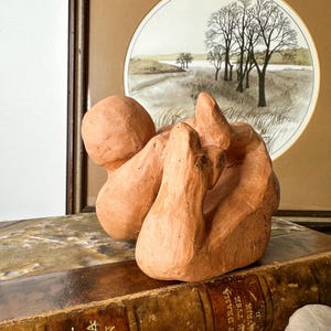 May include: A terracotta-colored abstract sculpture with rounded forms. The sculpture sits atop a stack of antique books with gold lettering. A framed landscape painting with trees and a field is in the background.