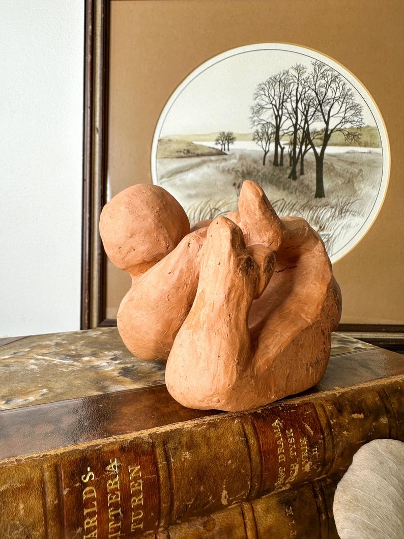 May include: A terracotta-colored abstract sculpture with rounded forms. The sculpture sits atop a stack of antique books with gold lettering. A framed landscape painting with trees and a field is in the background.