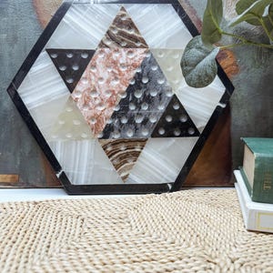 May include: A hexagonal Chinese checkers board crafted from polished stone. The board features a central star pattern composed of triangular sections in white, black, brown, and reddish-brown marble, each containing small circular indentations for game pieces.