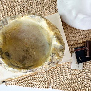 May include: A decorative, irregularly shaped bowl with a light yellow and gray color scheme. The bowl sits on a stack of books, with a woven mat in the background. Small matchboxes are also visible.