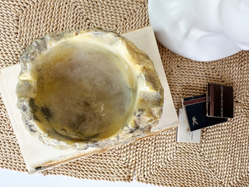 May include: A decorative, irregularly shaped bowl with a light yellow and gray color scheme. The bowl sits on a stack of books, with a woven mat in the background. Small matchboxes are also visible.