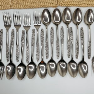 May include: A collection of stainless steel flatware including forks and spoons with a textured floral pattern on the handles. The set consists of various sizes of spoons and forks arranged in rows, featuring polished metal bowls and tines.